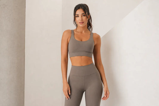 Mulher sorrindo, vestindo conjunto de top e legging de cintura alta em tons neutros, em um estúdio clean com luz suave.